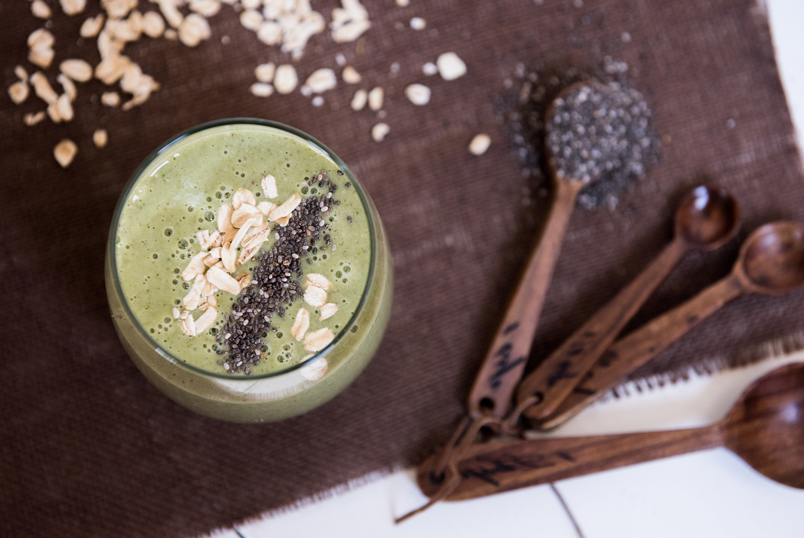 All Day Energy Smoothie in a clear glass, topped with oats and chia seeds, with scattered oats and chia seeds, next to wooden spoons with chia seeds.