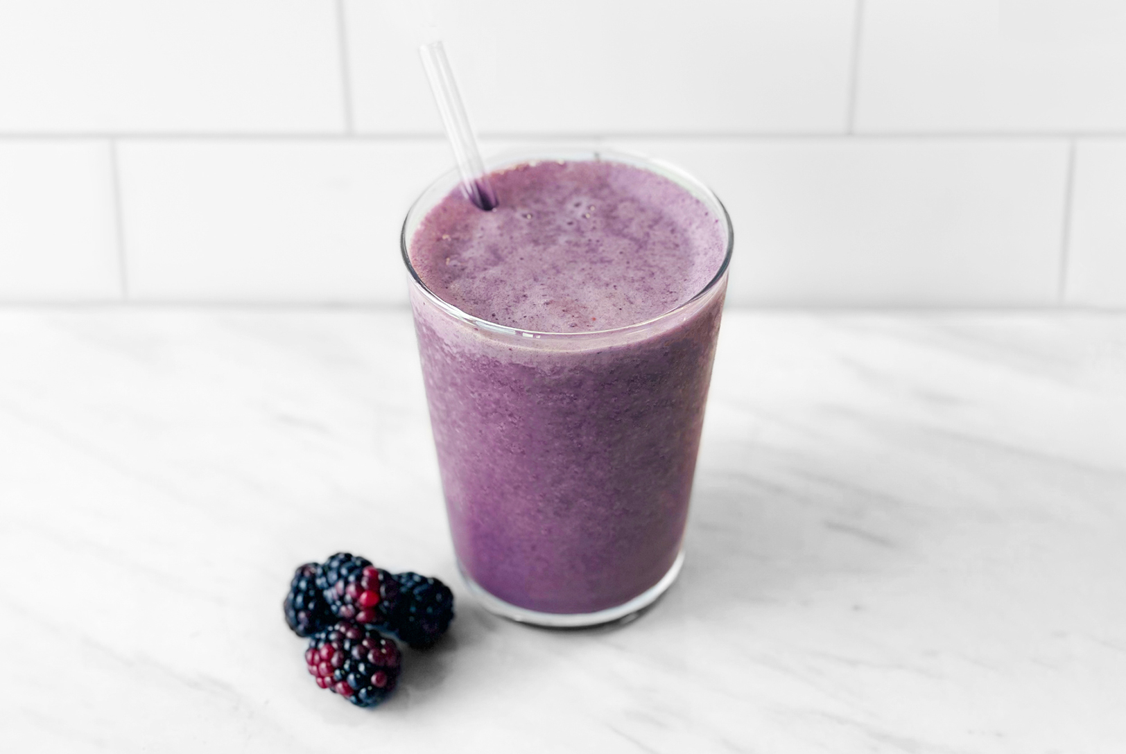 A tall glass filled with a purple blackberry smoothie with a clear straw, next to fresh blackberries on a white countertop.