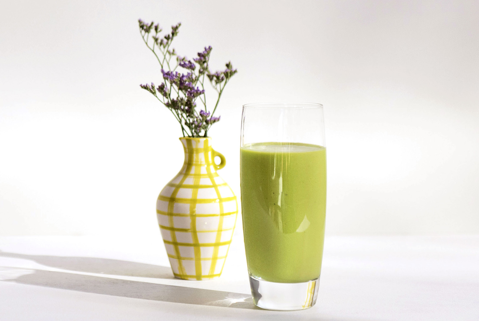 Creamy green Pineapple Delight Smoothie in a tall glass next to a decorative yellow and white vase with delicate purple flowers.