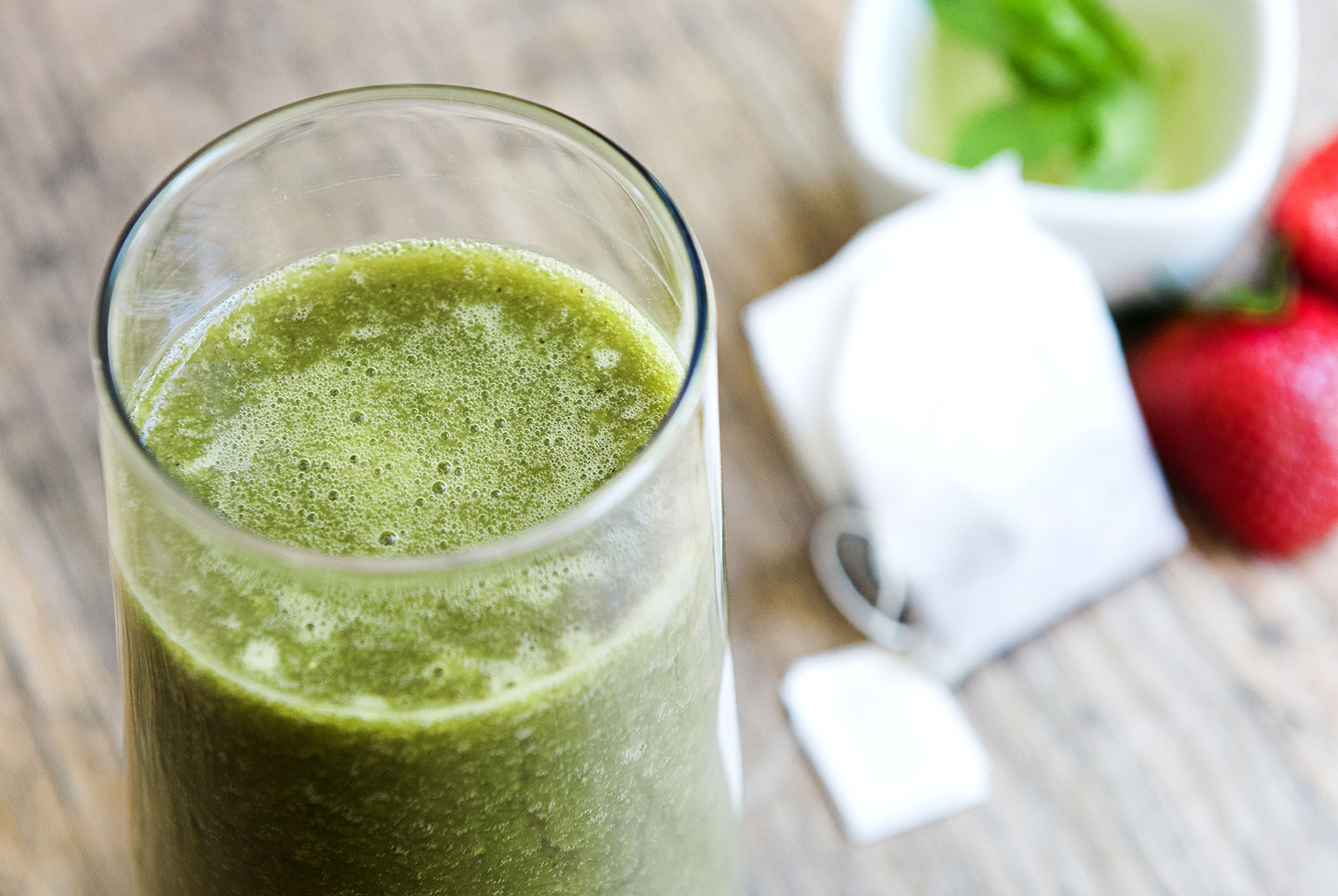 green smoothie in a glass next to green tea, fresh strawberries, and fresh mint leaves 