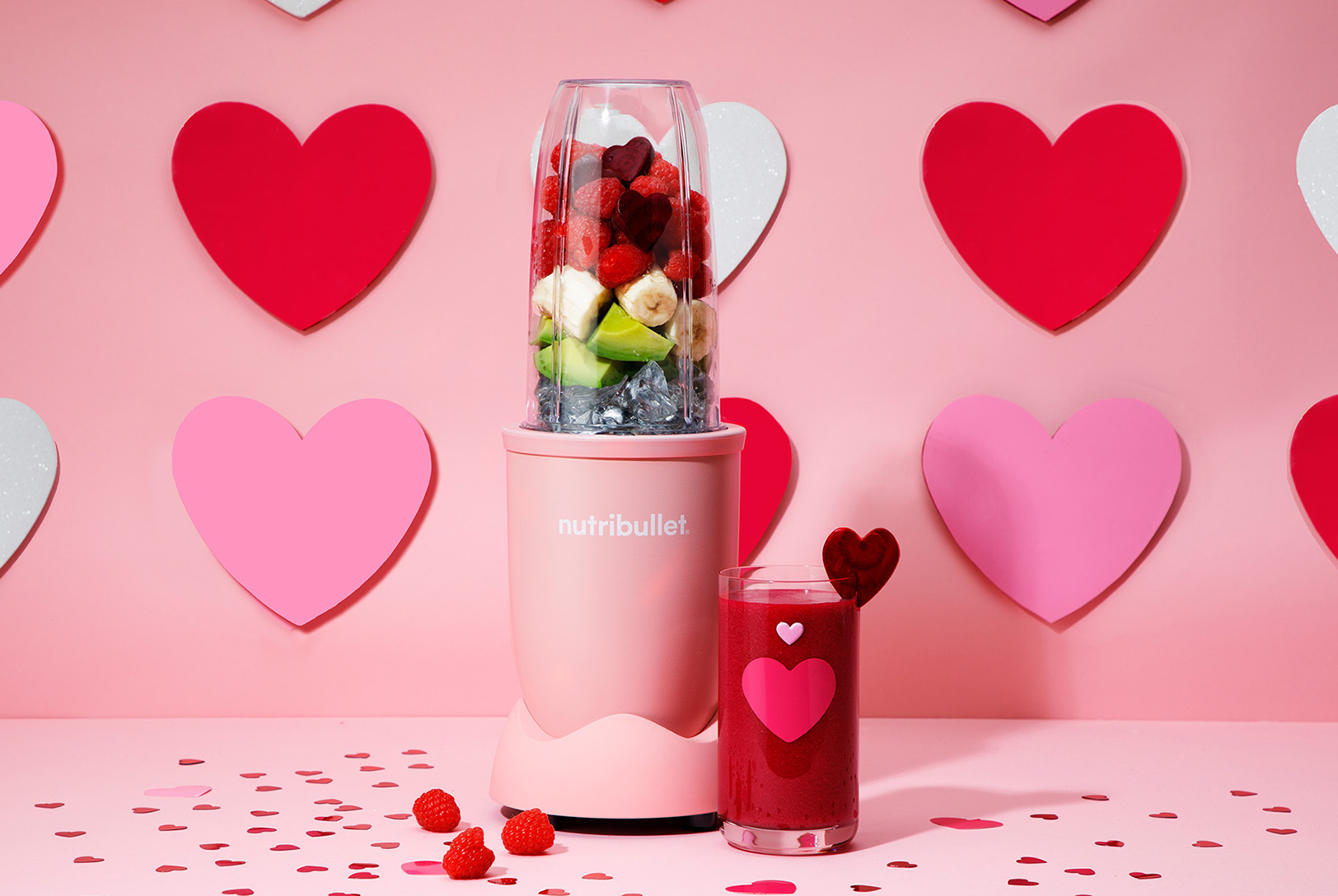Pink nutribullet single serve blender with fruits with a cup of red beet smoothie in front of a wall of hearts