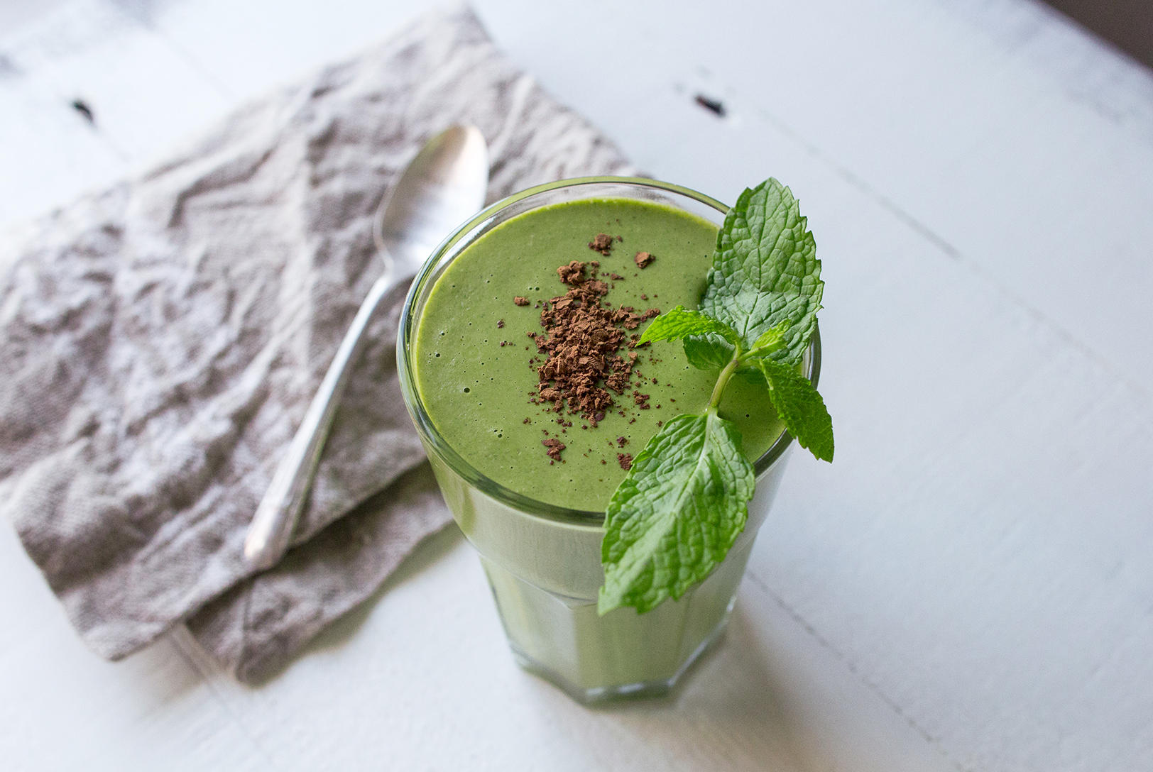 Mint Condition Smoothie in a glass, topped with chocolate shavings and fresh mint leaves, on a textured white surface with a napkin and spoon.