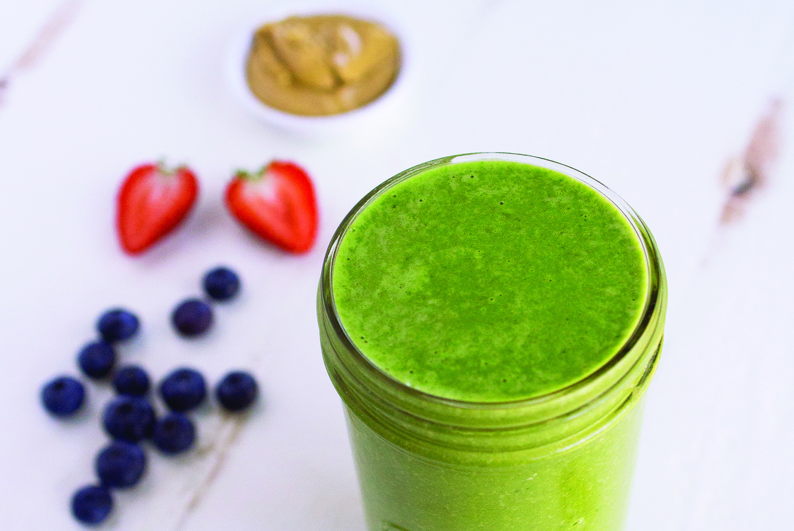 Vibrant green PB & J Smoothie in a glass jar, surrounded by fresh blueberries, sliced strawberries, and peanut butter.