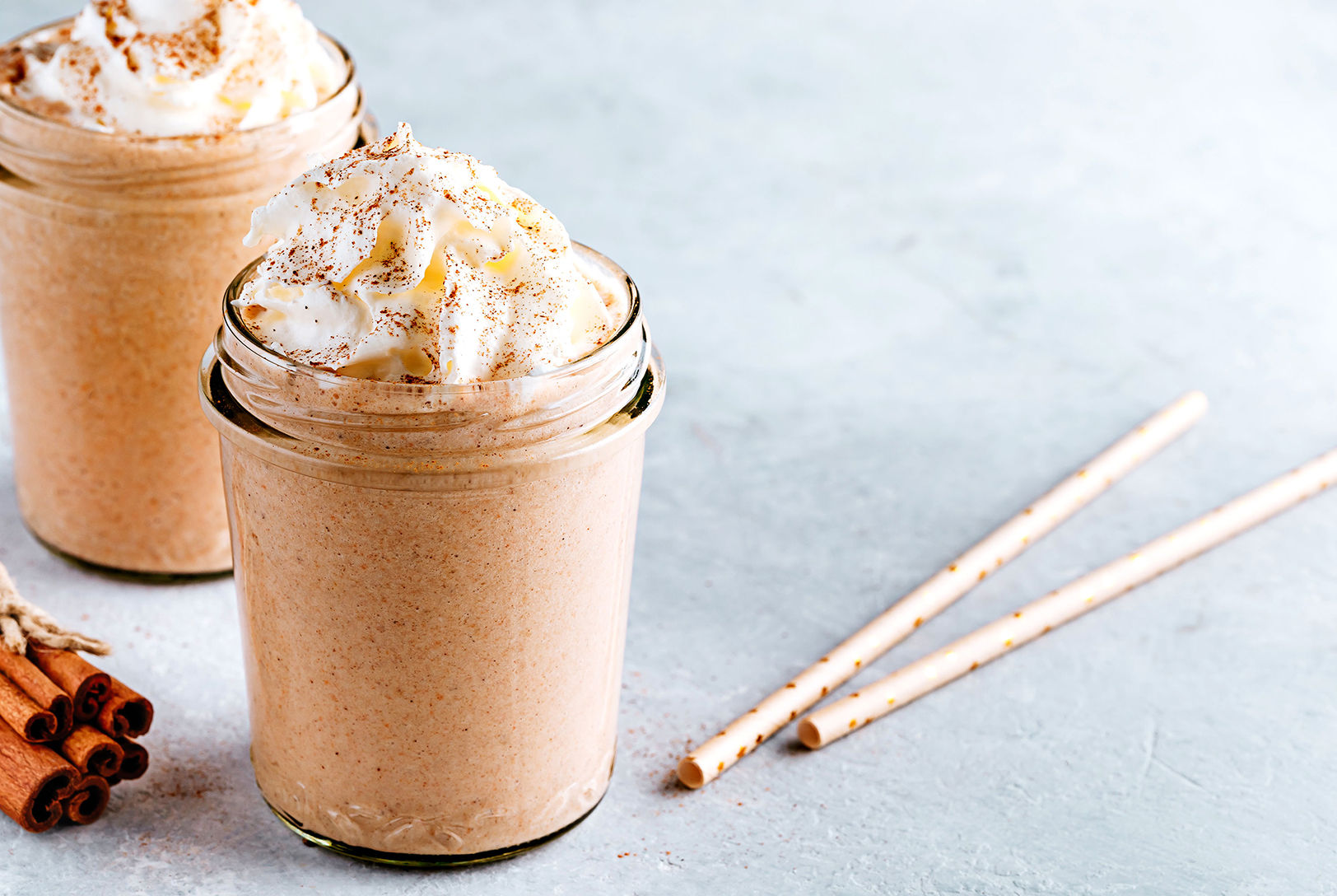 Creamy pumpkin spice smoothies in glass jars with whipped cream and spice, accompanied by cinnamon sticks and straws.