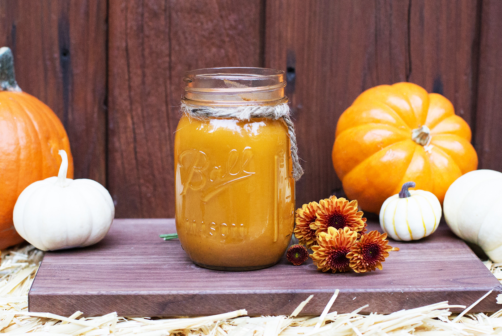 Pumpkin banana smoothie in a mason jar on a wooden board with small pumpkins and orange flowers.