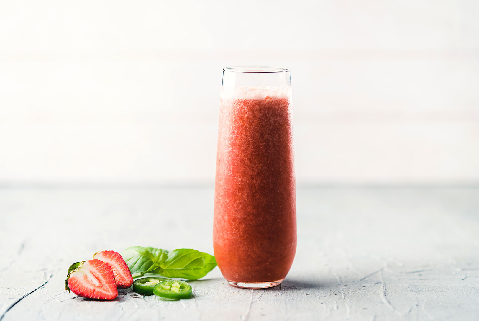 A tall glass with Strawberry Basil and Jalapeño Essence Water, next to sliced strawberries, basil leaves, and sliced jalapeños.