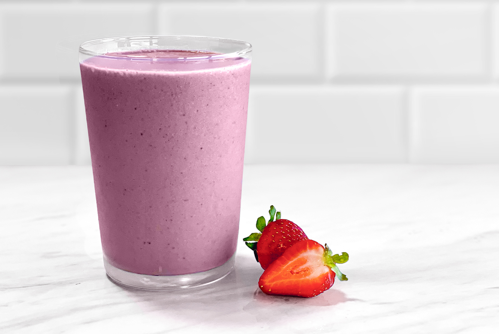Glass with light purple Strawberry Protein Punch smoothie, next to fresh strawberries on a white marble countertop.
