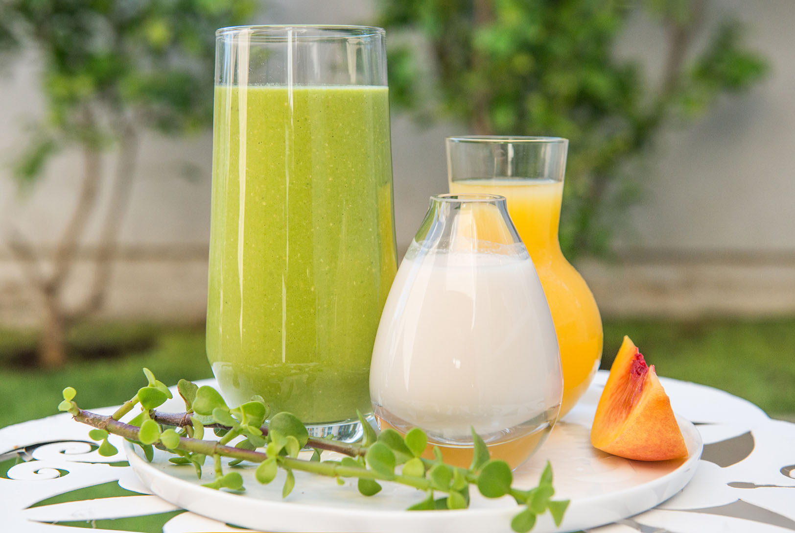 A tall glass of Vegan Peach Oat Smoothie on a tray, with a small carafe of oat milk, a glass of peach juice, and a slice of peach, garnished with a sprig.