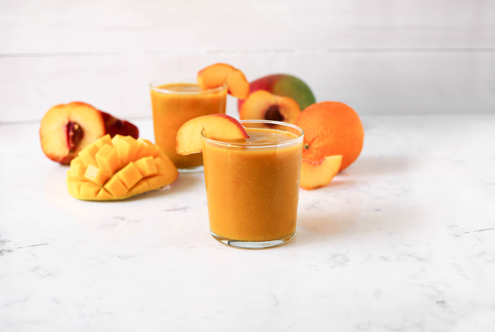 Two glasses filled with Orange Peach Mango Smoothie, with peach slices, surrounded by fresh peaches, an orange and a carved mango on a white surface.