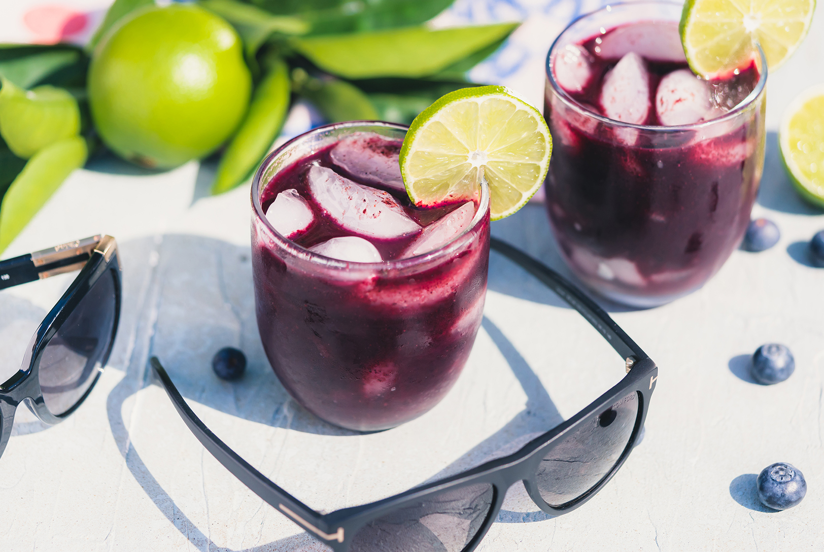 Berry Skinny Cocktail in two glasses with ice and lime slices, surrounded by fresh blueberries and sunglasses in a bright, summery setting.