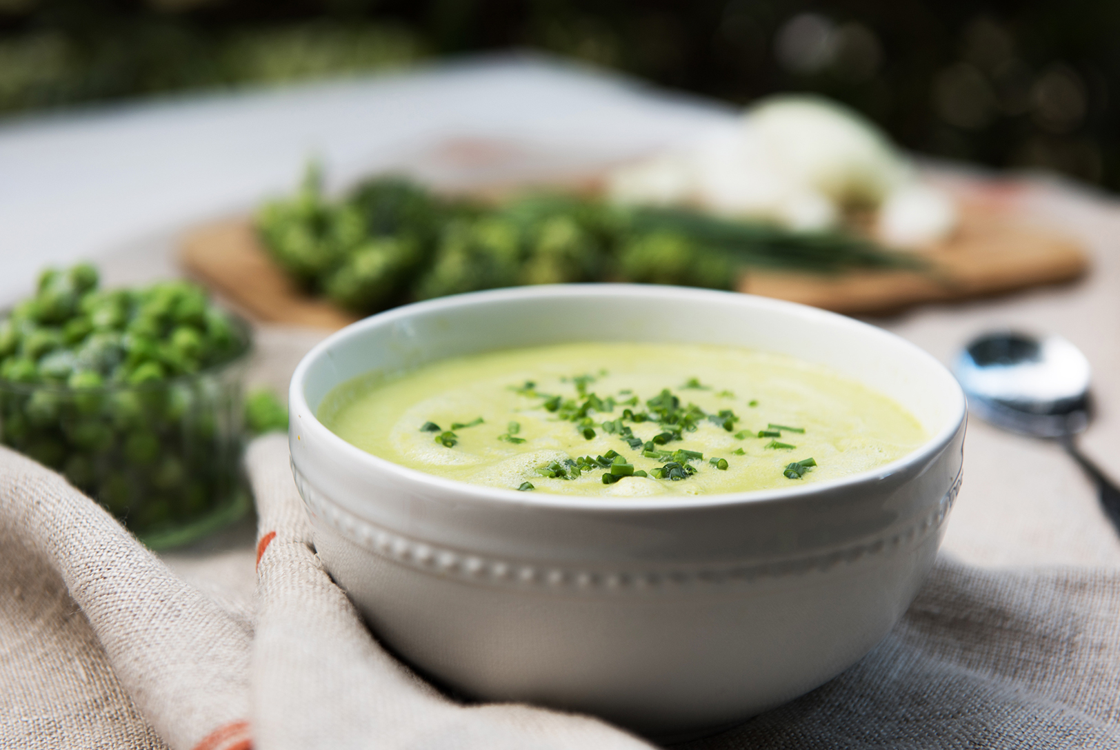 A bowl with green Broccoli, Pea, and Coconut Soup, sprinkled with freshly chopped herbs, a small glass bowl of bright green peas sits nearby.