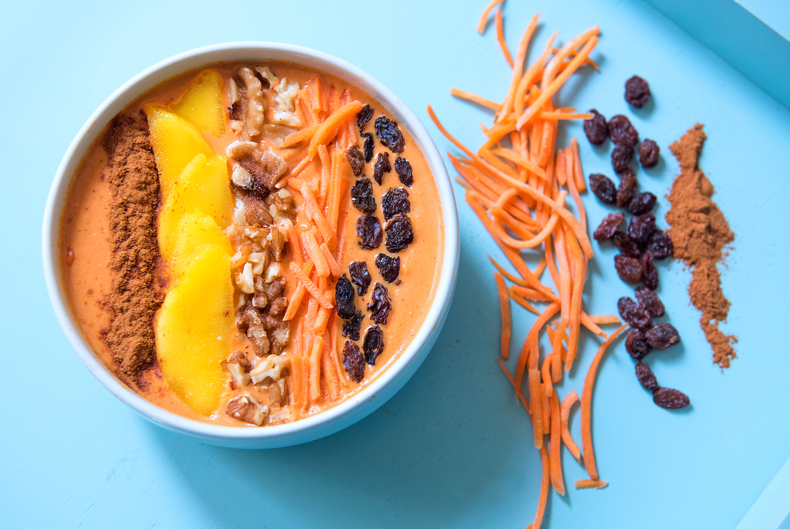 Carrot Mango Smoothie Bowl topped with mango, shredded carrots, walnuts, and raisins, with shredded carrots, raisins and cinnamon powder beside it.