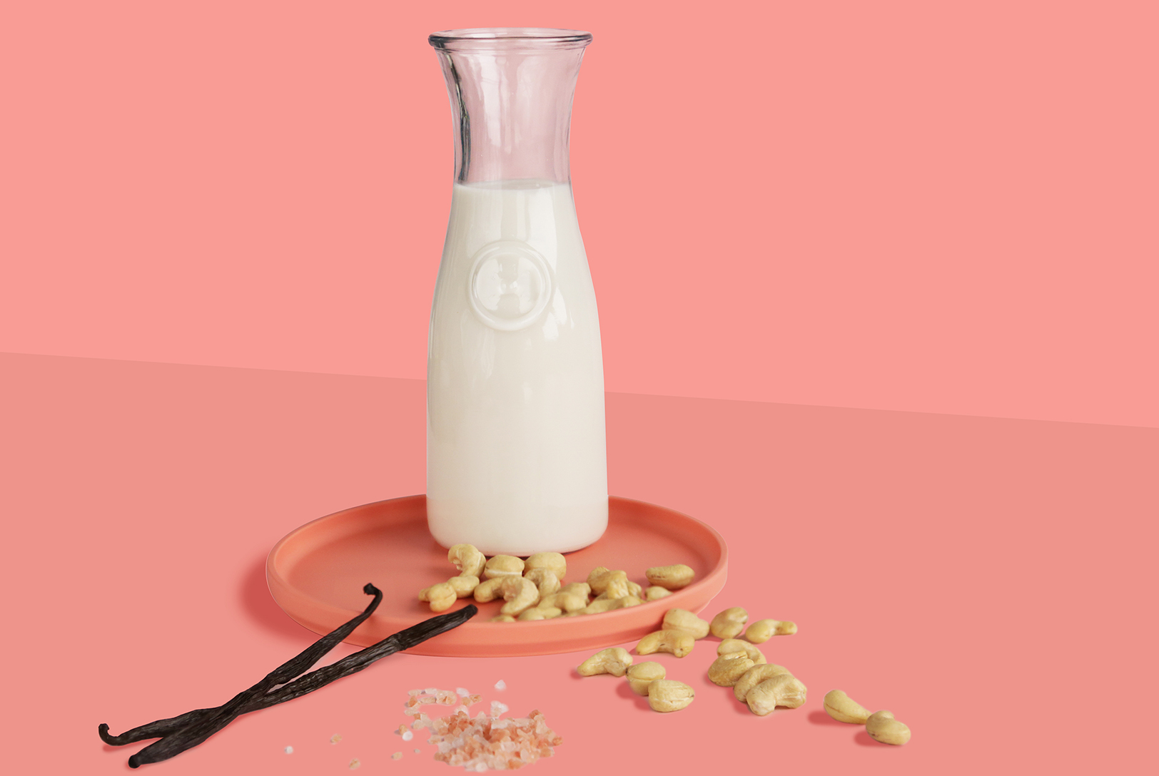 Cashew Milk in a glass carafe on a pink plate, surrounded by raw cashews, vanilla beans, and a sprinkle of salt on a soft pink background.