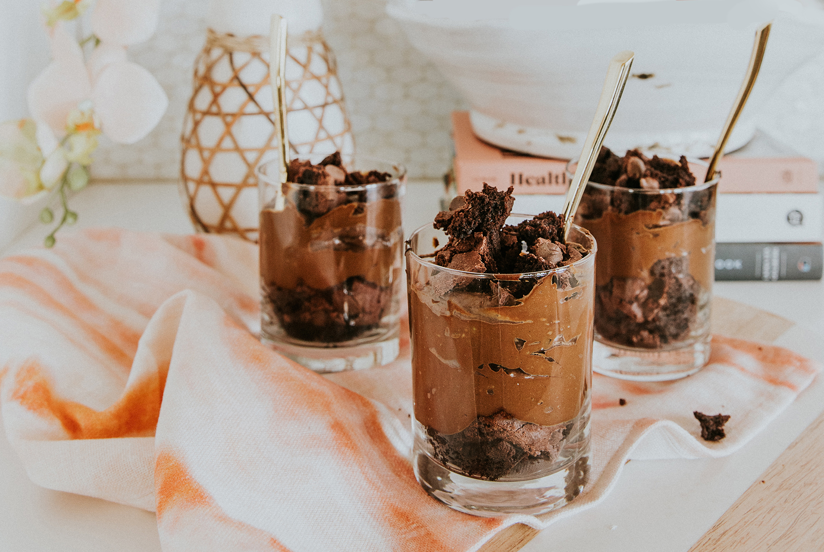Three chocolate avocado pudding in small glasses topped with brownie pieces with a stylized background.