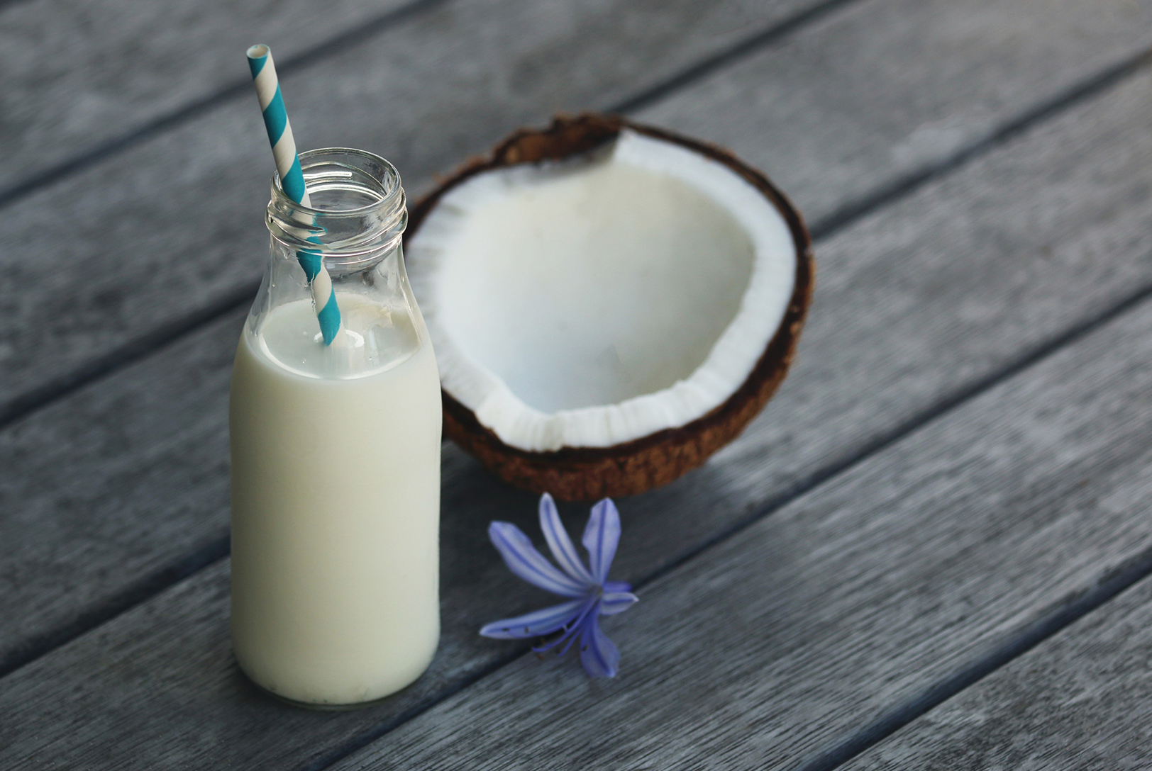 A small glass bottle with Coconut Milk with a striped straw, next to a halved coconut and a purple flower on a wooden surface.