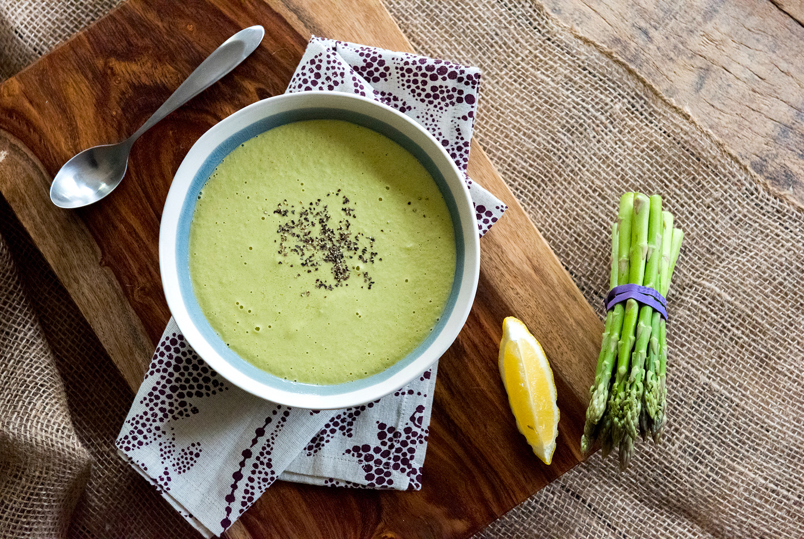 A bowl of light green Cream of Asparagus Soup, lightly seasoned, alongside fresh asparagus, a bright lemon wedge, and a silver spoon on a wooden board.
