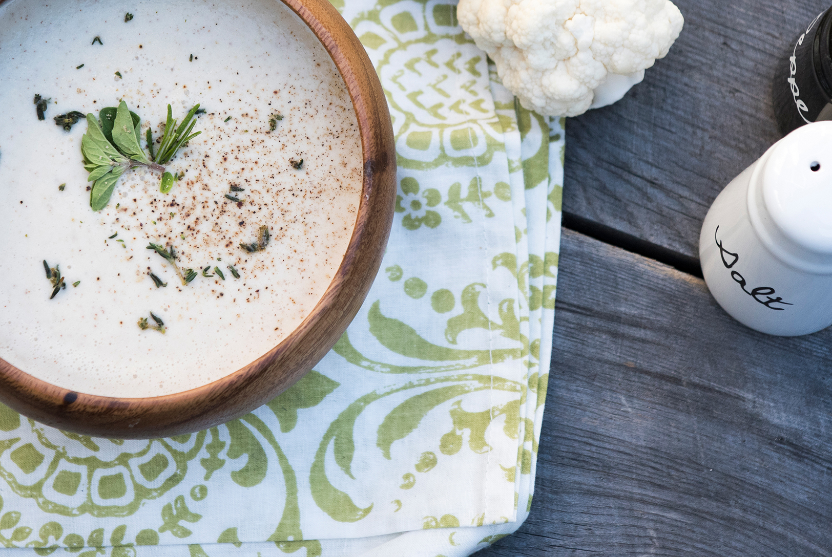 Creamy Cauliflower Soup in a wooden bowl, garnished with herbs and a sprinkle of spices, on a green patterned napkin, next to a head of cauliflower.