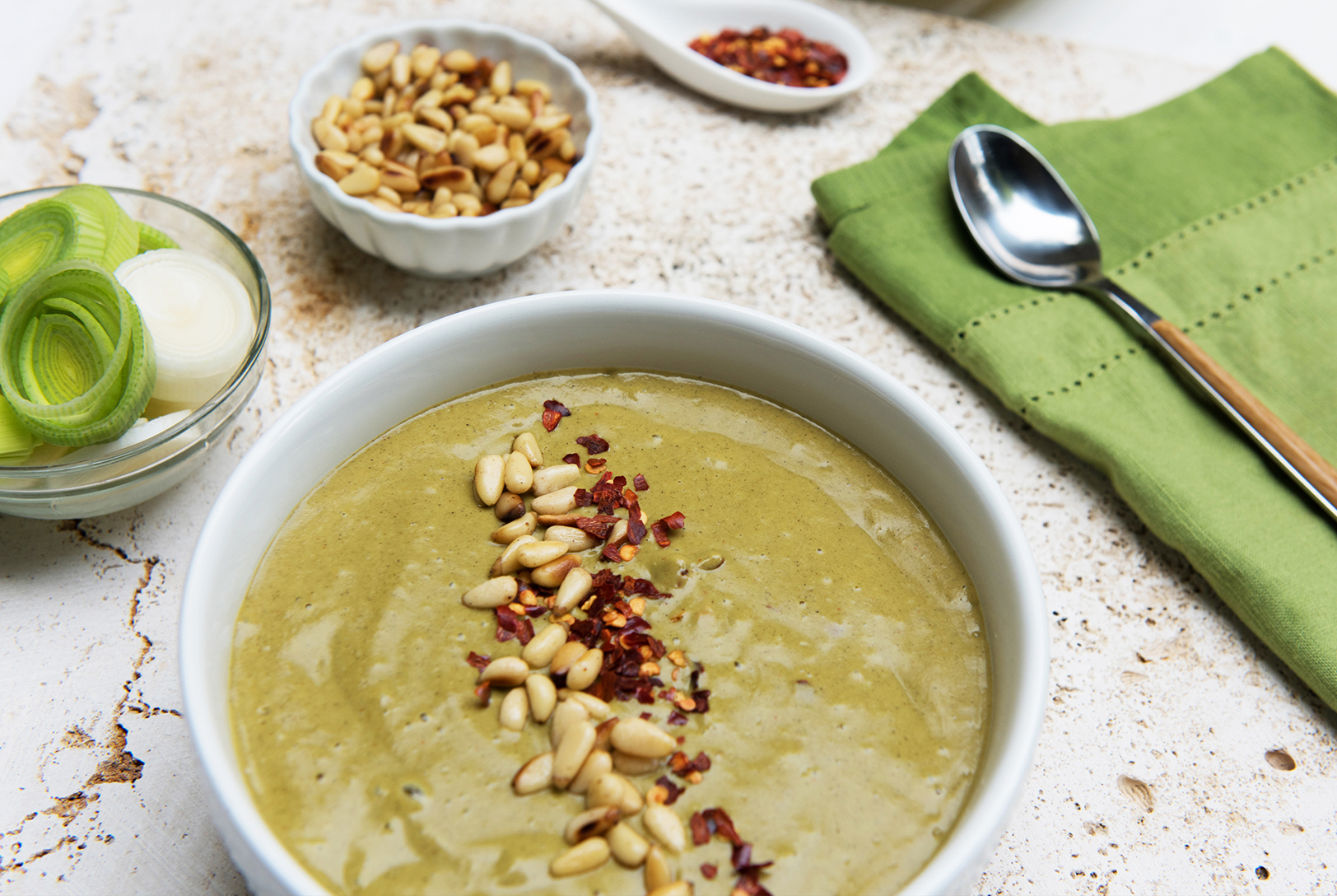 A bowl of creamy light green Roasted Leek and Zucchini Soup topped with pine nuts and red pepper flakes.