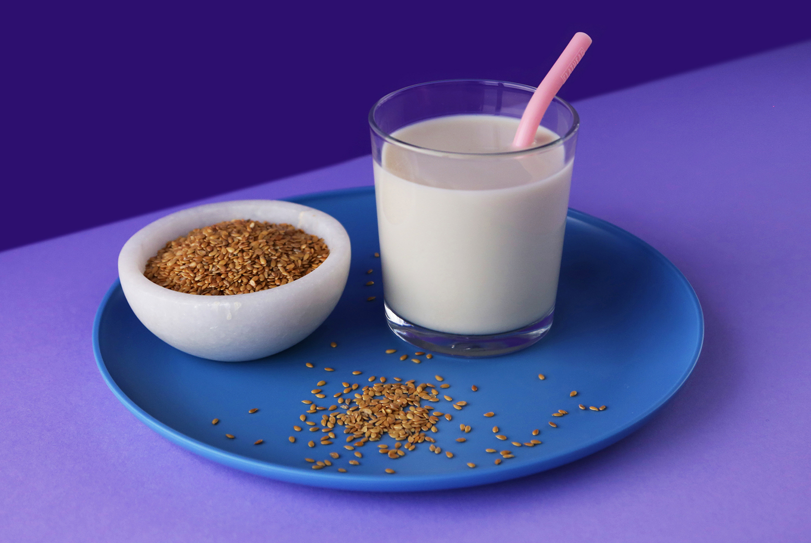 Glass of smooth Flax Milk with a pink straw, next to a bowl of flax seeds on a blue plate, with purple background.