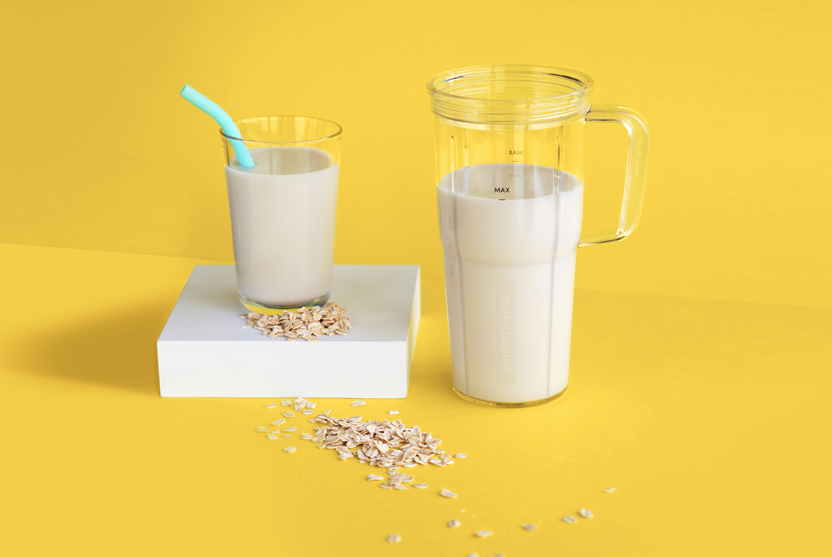 Gluten-Free Oat Milk shown in a drinking glass with a blue straw and in a blender cup, with scattered oats on a bright yellow surface.