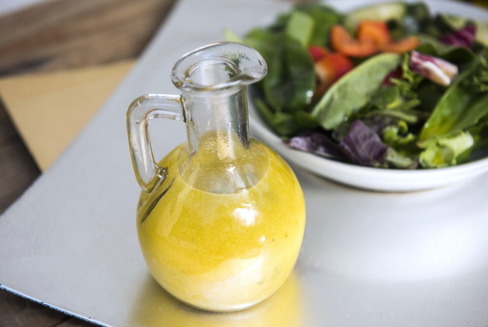 Greek Salad Vinaigrette in a glass cruet, with a salad in the background, over a silver surface.