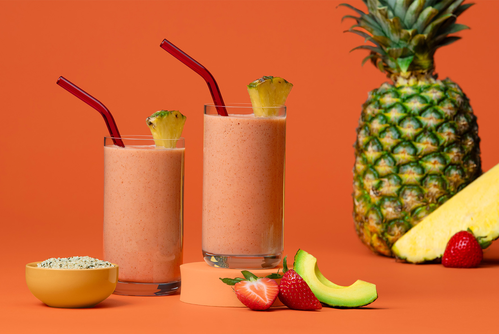 Two glasses of a Heart Healthy Smoothie with red straws and pineapple wedges, alongside pineapple, strawberries, avocado, and a bowl of seeds.