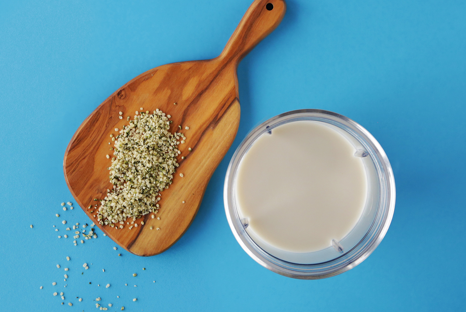 A glass of Hemp Seed Milk next to a wooden board with hemp seeds, on a bright blue background.