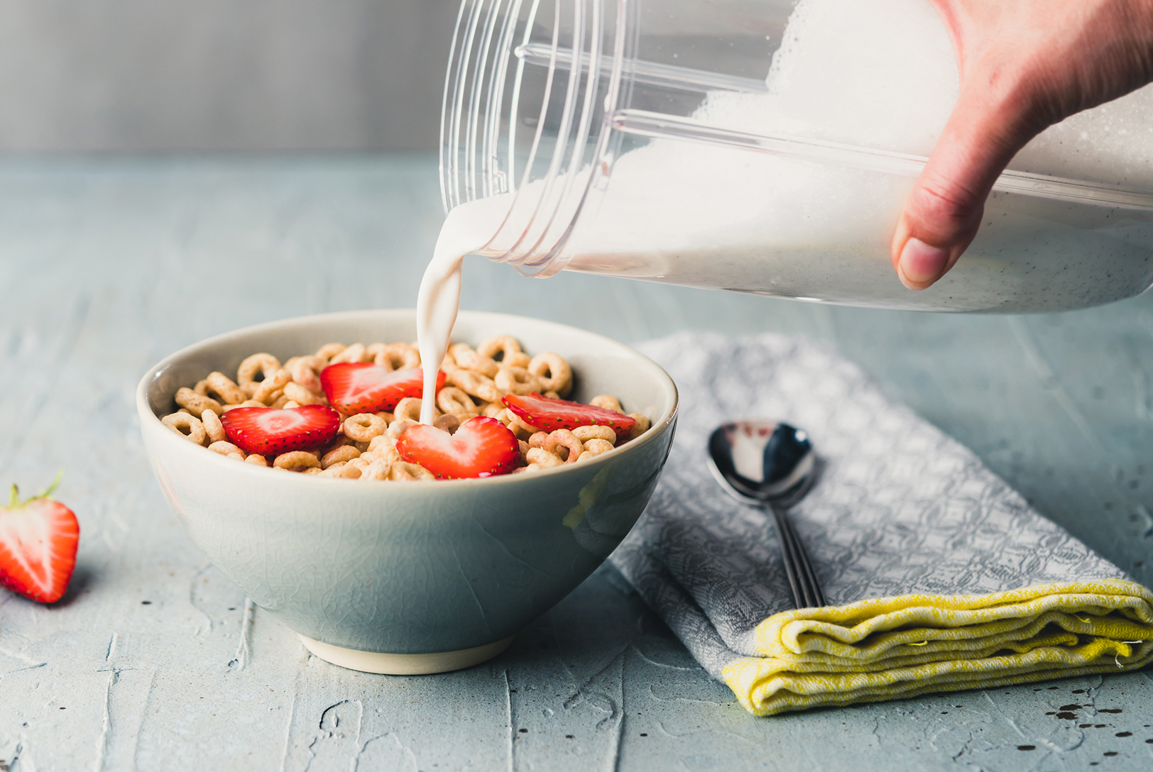 A bowl of cereal topped with sliced strawberries, with hemp milk being poured from a clear container, with a spoon and napkin.