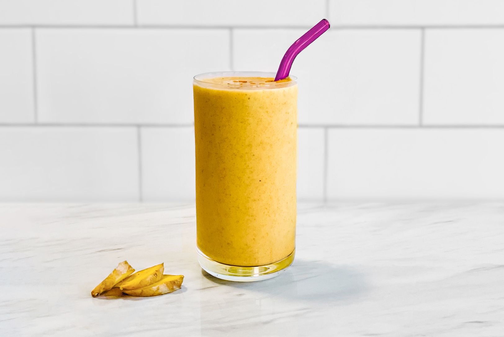 Ginger Mango Smoothie in a glass with a purple straw, next to fresh ginger pieces on a white marble countertop against a white tiled wall.