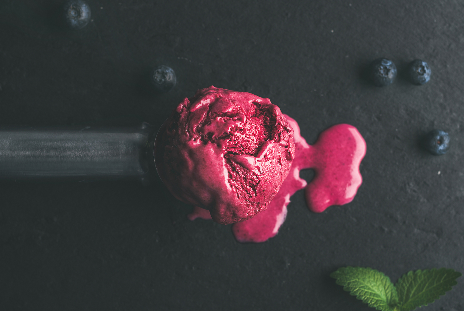 A scoop of pink "Make My Heart Beet" red velvet nice cream on a metal scoop, surrounded by blueberries and mint leaves, on a dark surface.