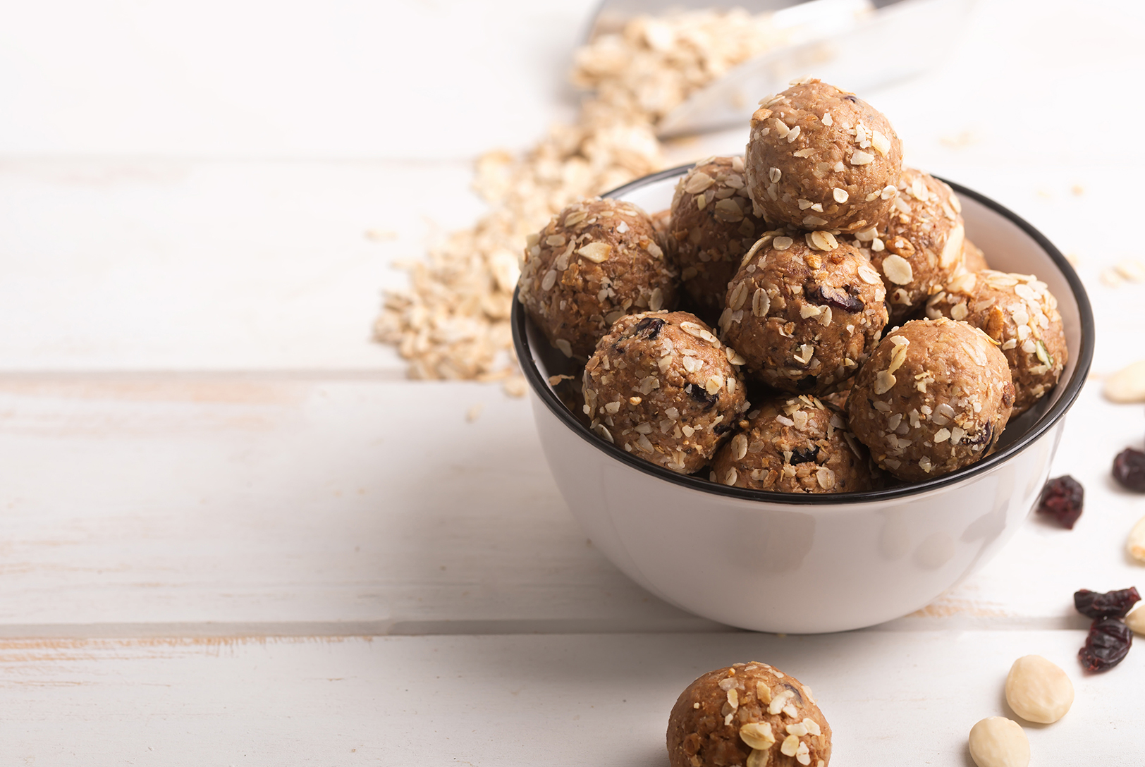 Peanut Butter Energy Bites with oats in a white bowl, surrounded with scattered oats and dried fruit on a white wooden surface.