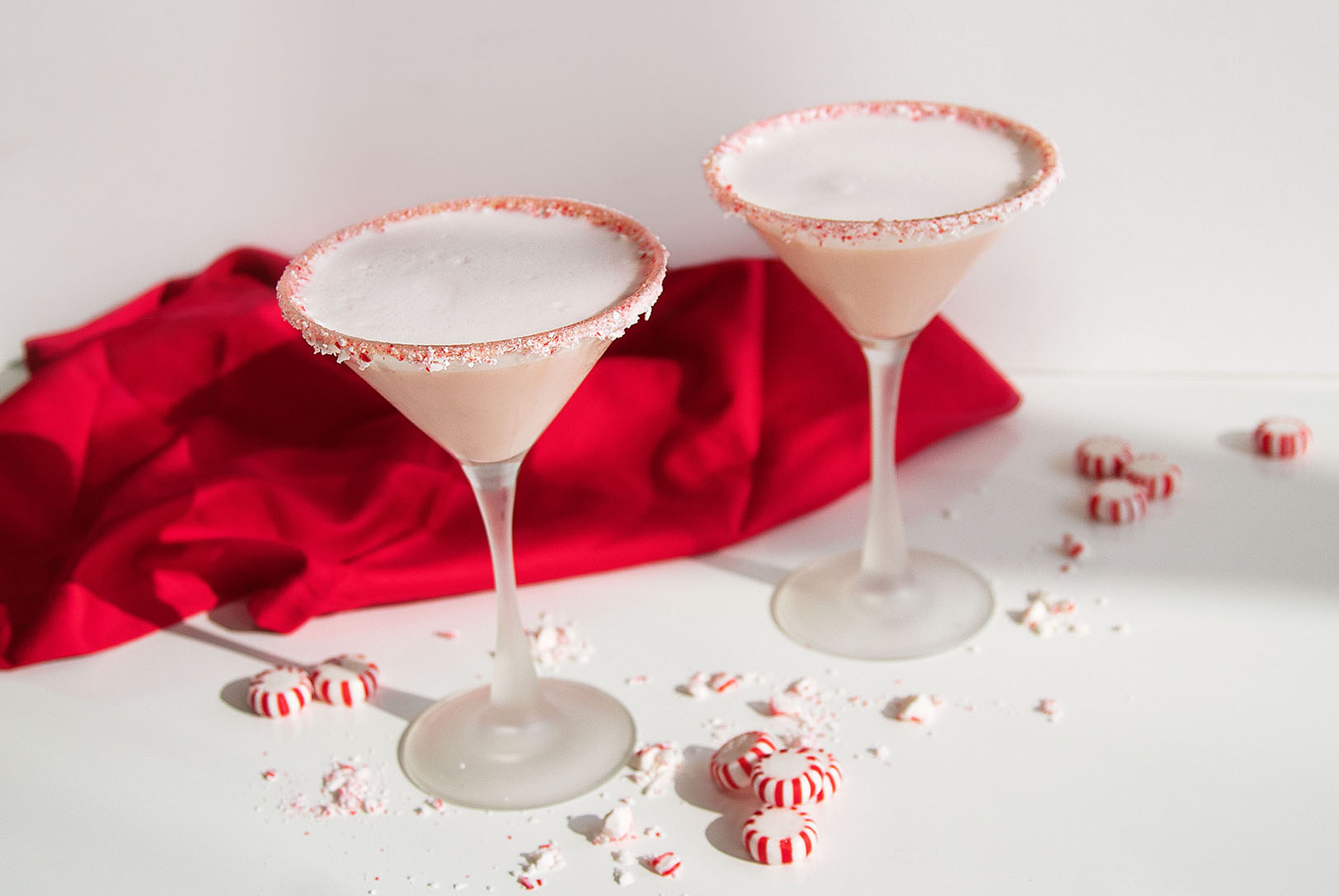 Festive Peppermint Martini cocktails in stemmed glasses with a crushed candy cane rim, set against a red cloth with scattered peppermints.