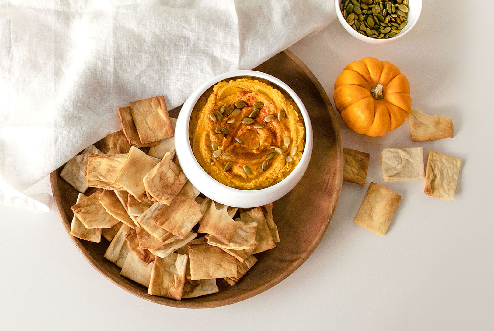 Creamy pumpkin hummus served in a bowl on a wooden platter, with crispy pita chips, a mini pumpkin, and a bowl of pumpkin seeds.