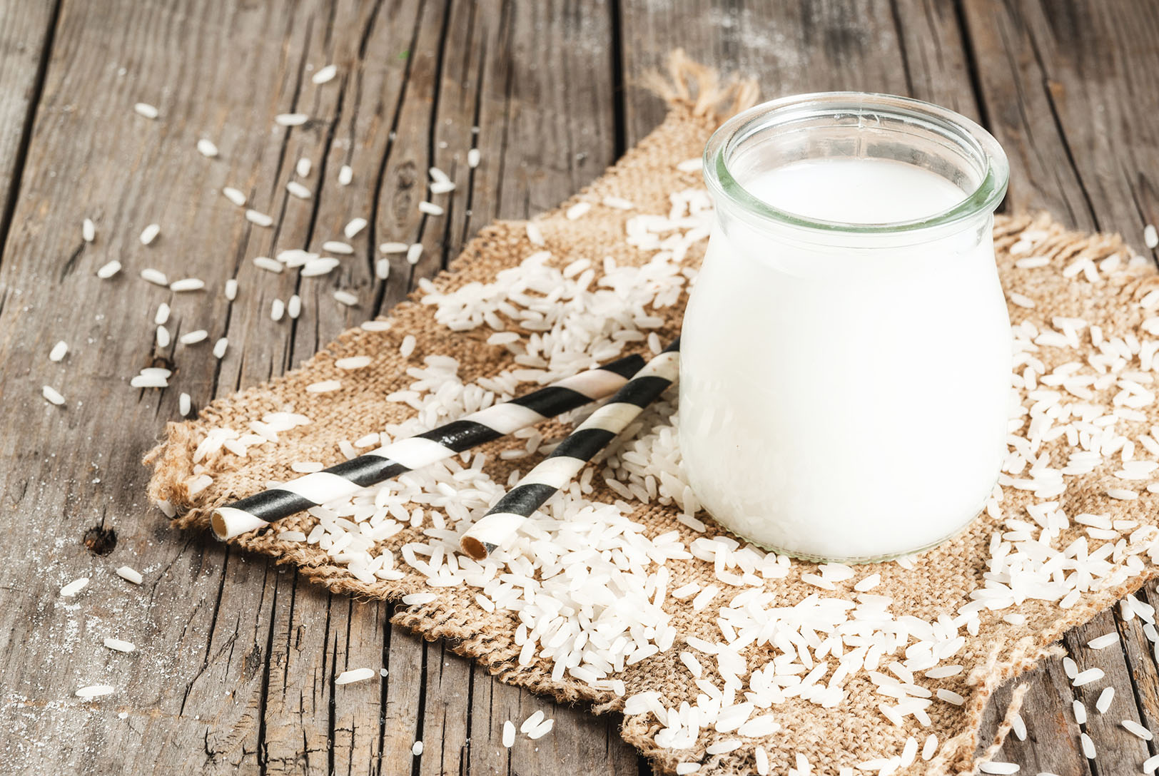 Rice Milk in a clear glass jar with black and white straws, sitting on a burlap square surrounded by scattered rice grains on a rustic wooden surface.