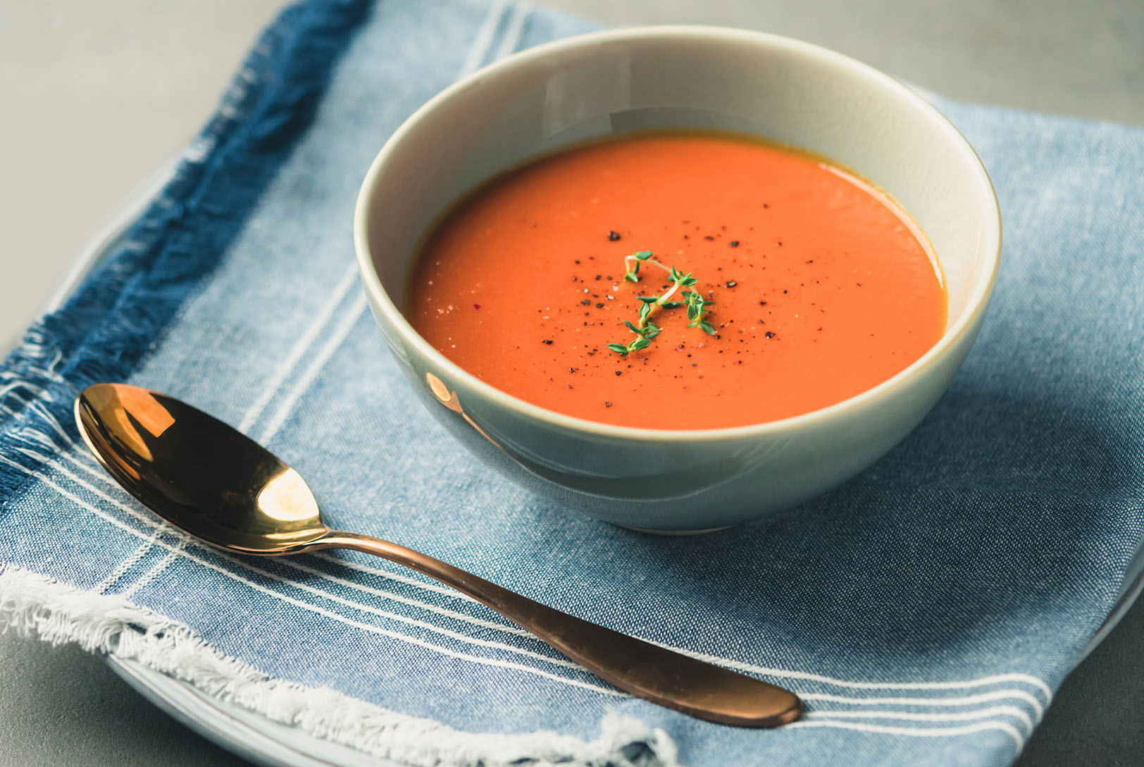 Roasted Red Pepper and Cauliflower Soup in a light green bowl, garnished with herbs and black pepper, sitting on a blue linen napkin with a gold spoon.