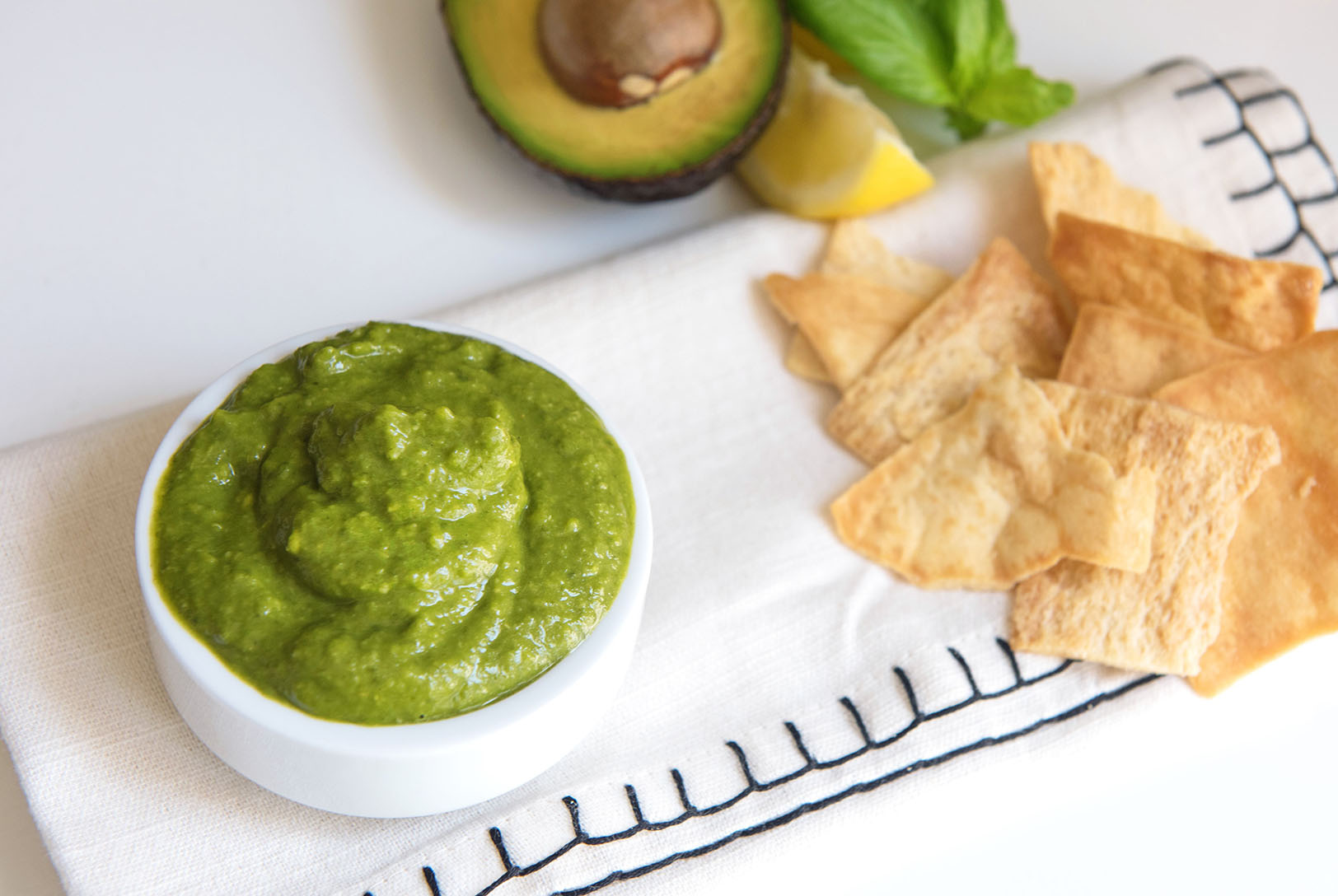 Spinach Avocado Pesto in a bowl, served with pita chips on a white napkin, with half an avocado, a lemon wedge, and basil leaves.