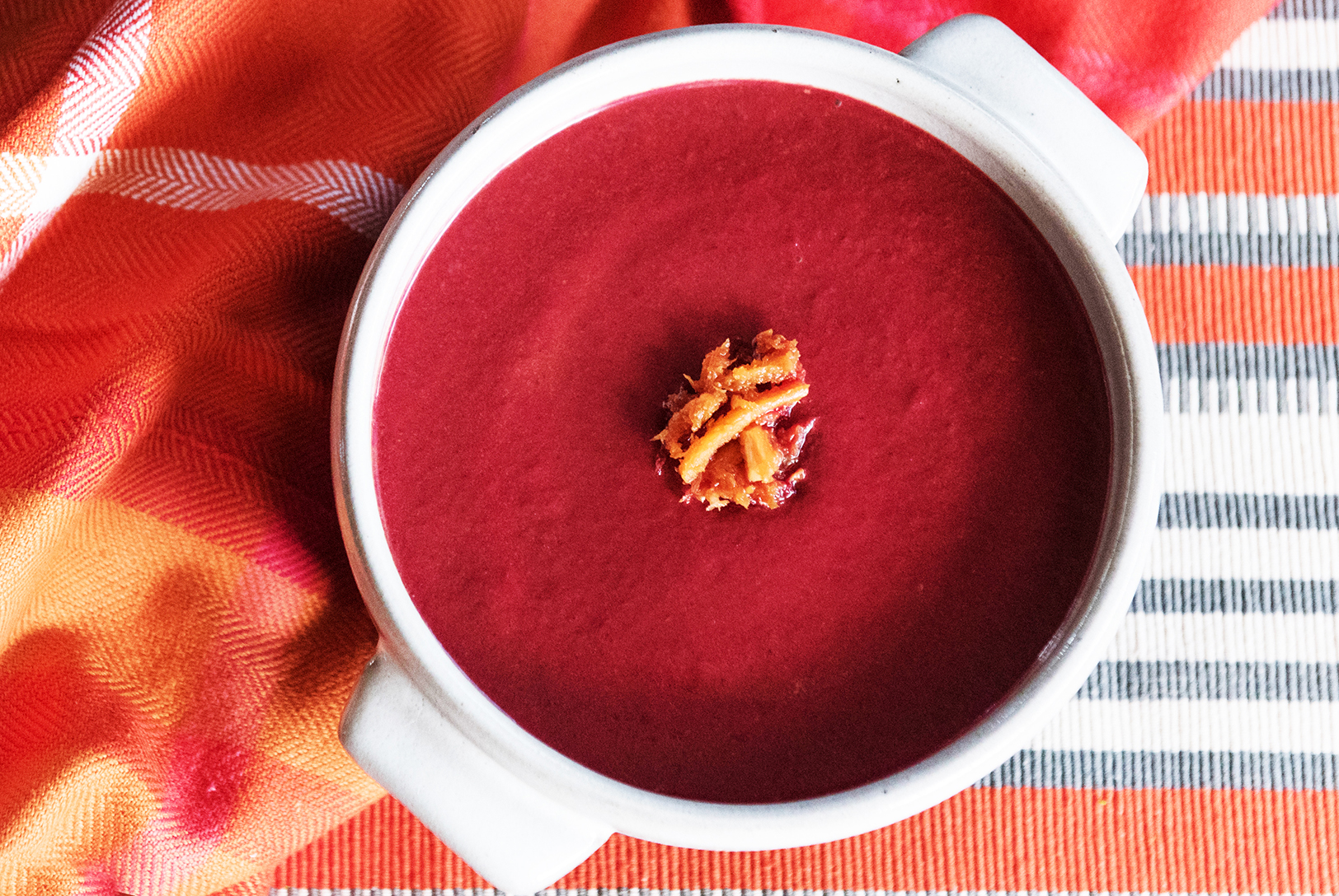 Vibrant crimson Superfood Root Soup in a white bowl, topped with a golden garnish, set against a colorful striped textile.