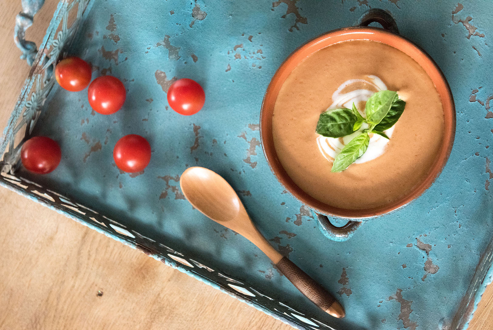 Tomato Basil Soup in a terracotta bowl, swirled with cream and garnished with fresh basil, on a rustic blue tray with cherry tomatoes and a wooden spoon.