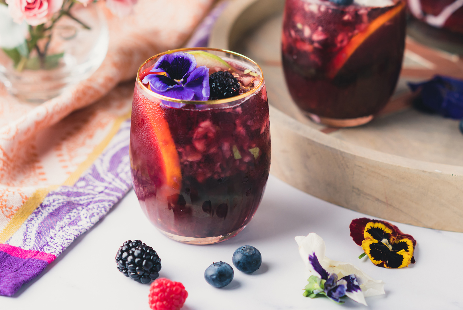 Two glasses of The Ultimate Sangria, filled with mixed berries and citrus slices, garnished with edible flowers on a light surface and wooden board.