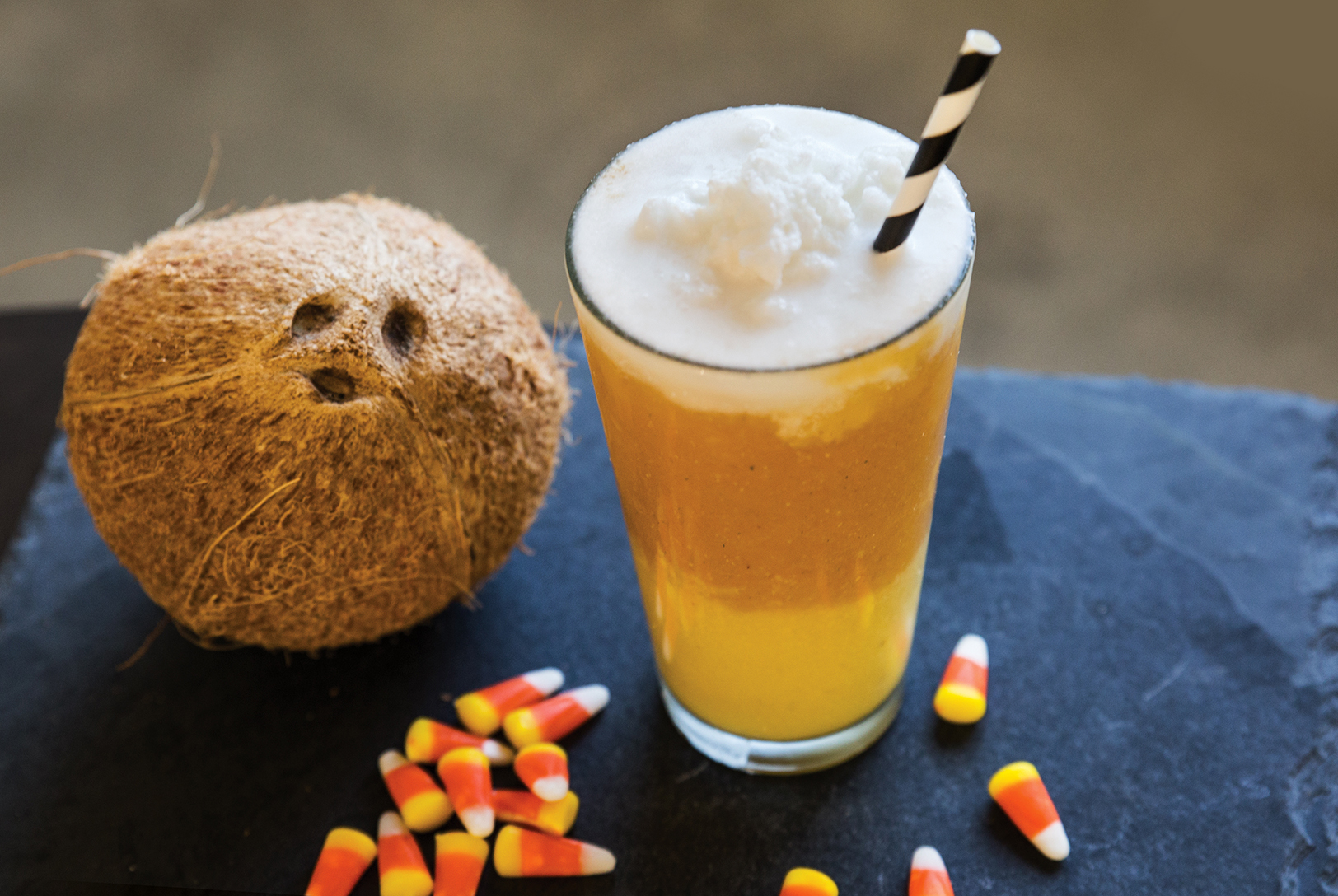 A yellow Tropical Candy Corn Smoothie in a glass with a black and white striped straw, next to a whole coconut and scattered candy corn pieces.