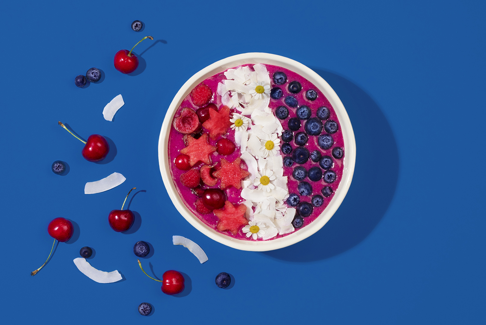 Festive Patriotic Smoothie Bowl, topped with star-shaped watermelon, coconut flakes, blueberries, cherries, and edible flowers on a bright blue background.