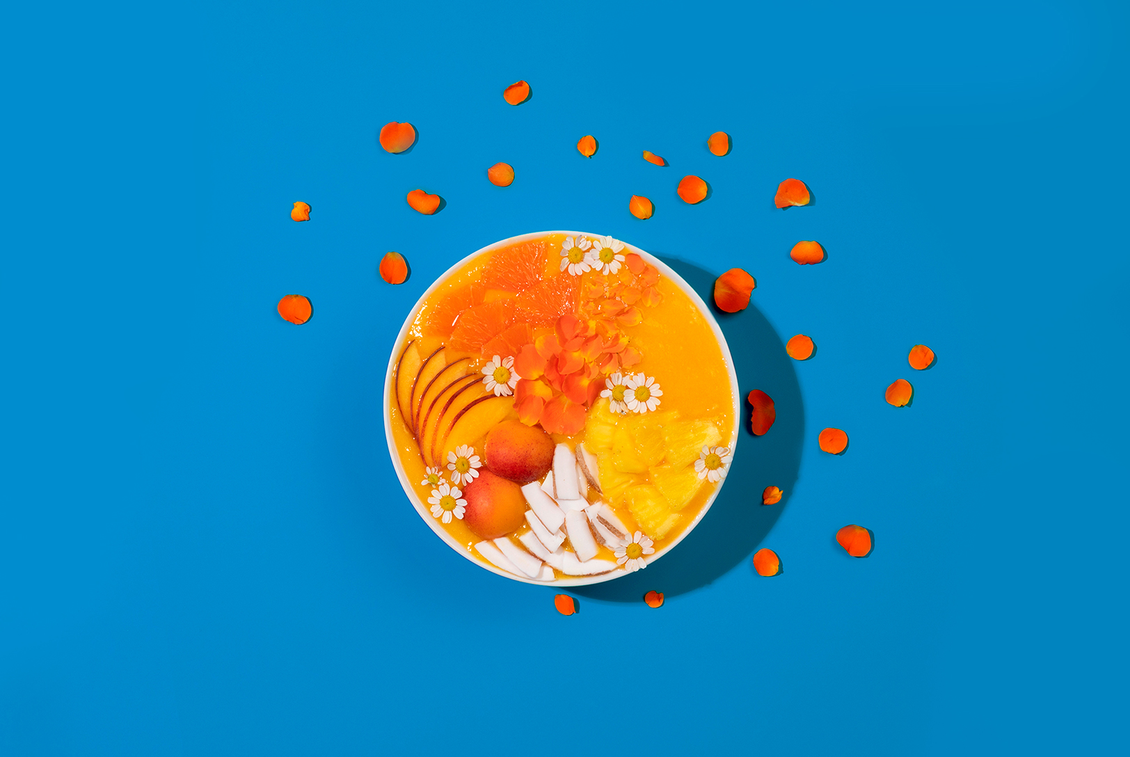 A Peach Smoothie Bowl on a blue background, topped with peaches, almonds, and white flowers, surrounded by scattered orange flower petals.