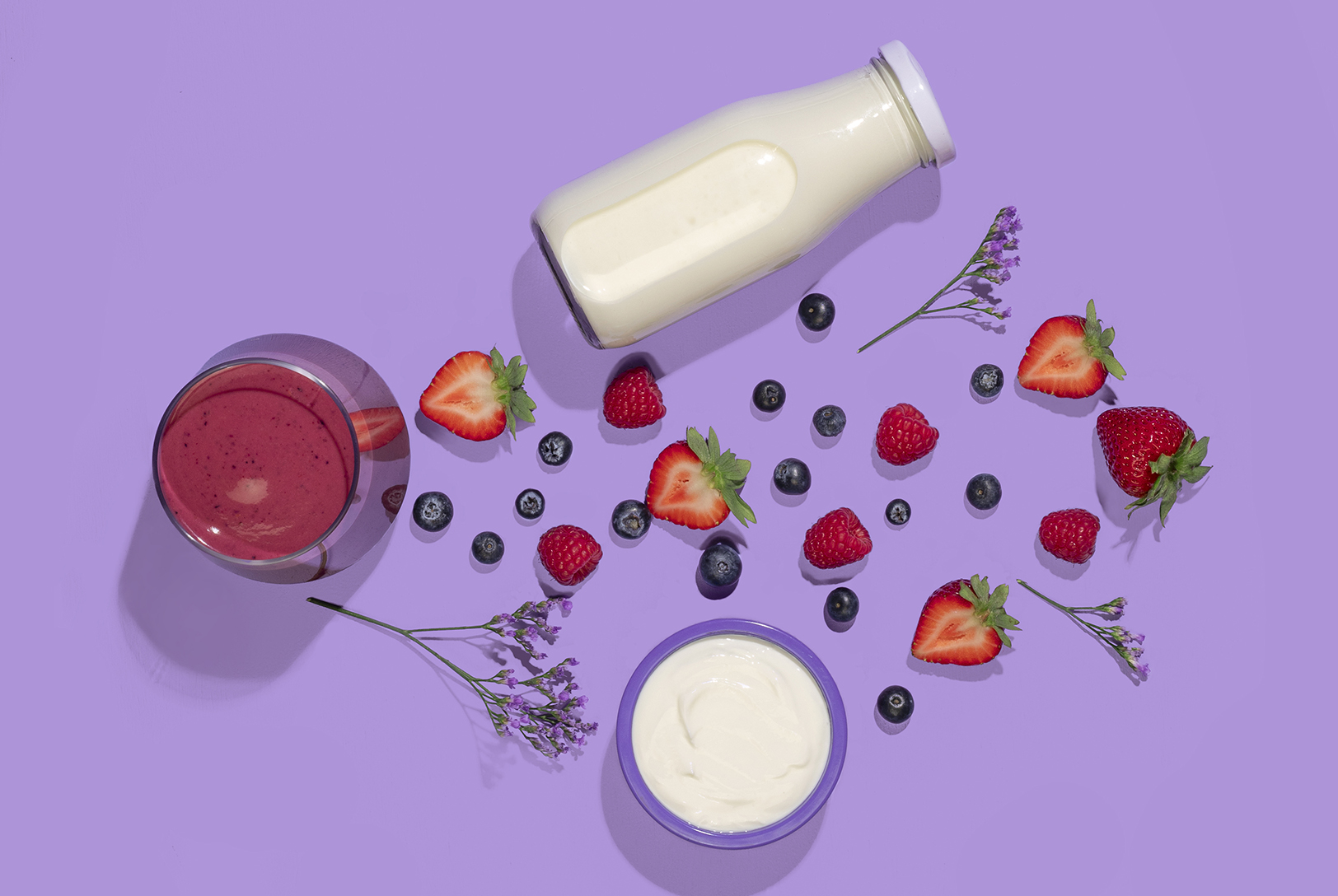 Overhead view of a Triple Berry Smoothie in a glass, alongside strawberries, raspberries, blueberries, almond milk, and yogurt on a purple background.