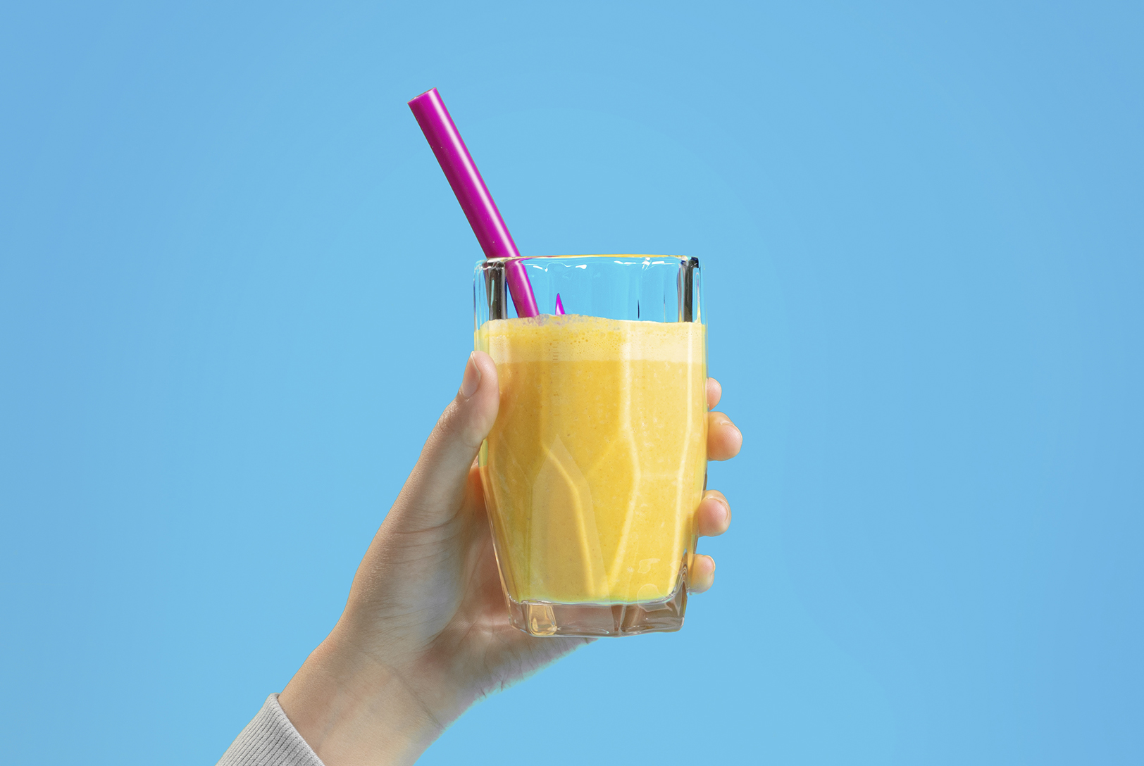 A hand holds a bright yellow Tropical Smoothie in a glass with a pink straw against a blue background.