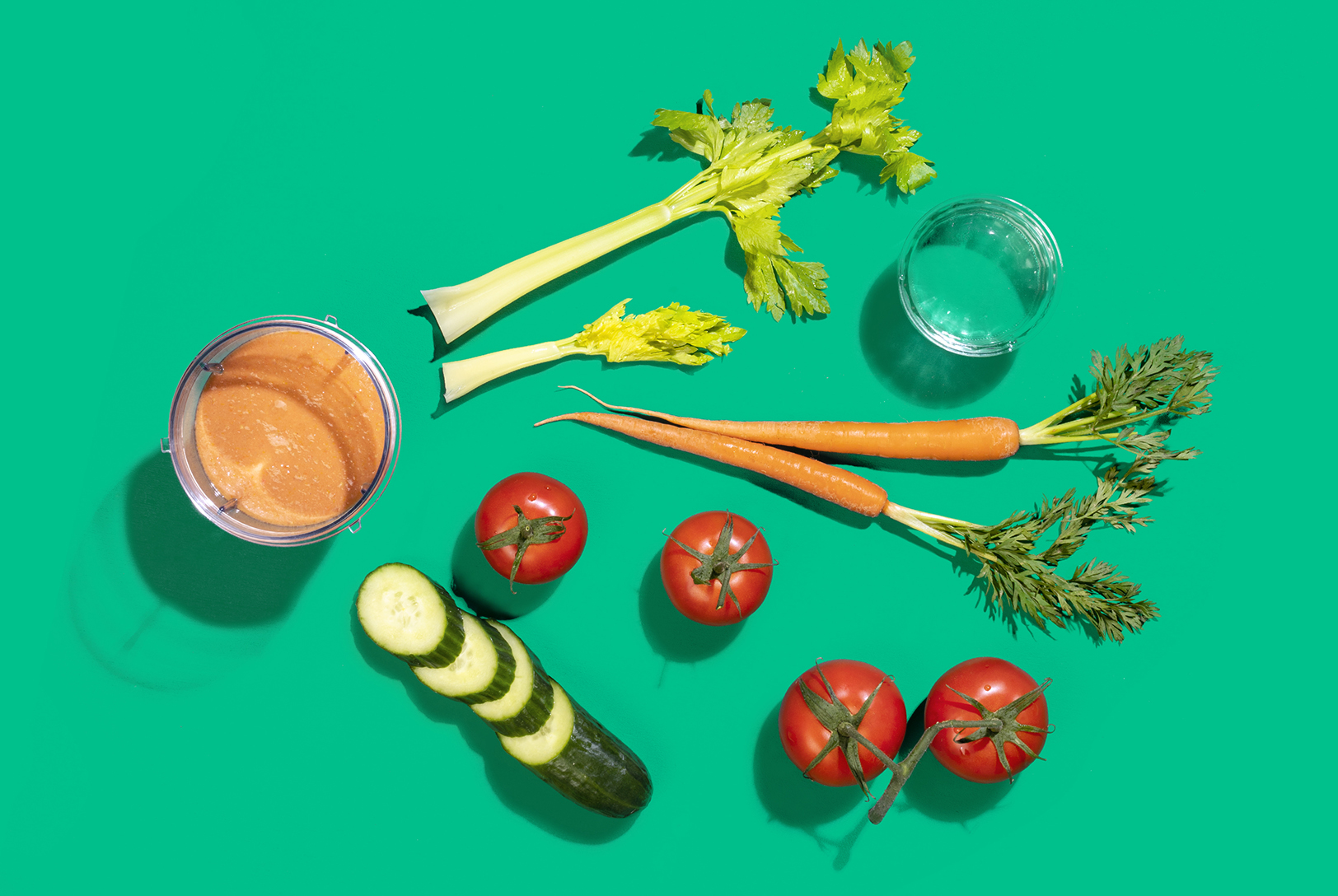 A top-down view of carrots, tomatoes, cucumber, celery stalks, and a glass of water arranged around a orange smoothie on a green background.