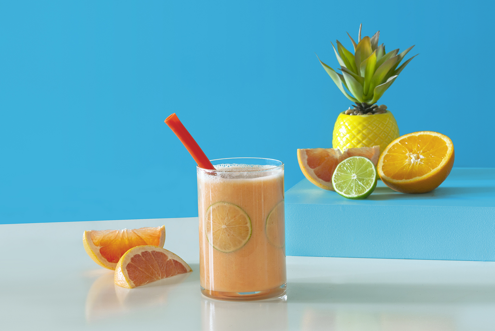 Orange citrus smoothie in a glass with red straw surrounded by sliced citrus fruits.