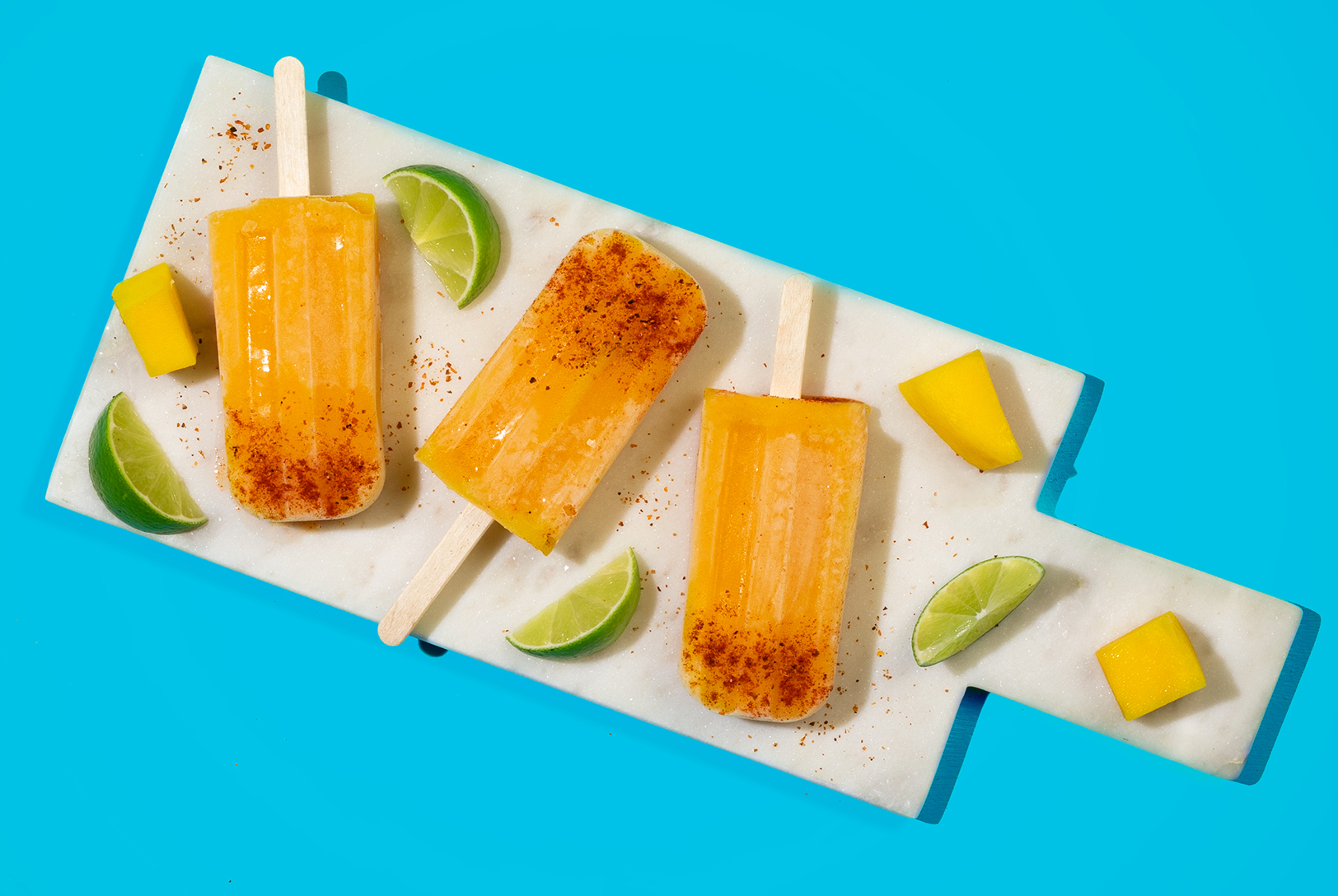 Three Mango Popsicles sprinkled with chili powder, with fresh mango chunks and lime wedges on a white marble board against a blue background.