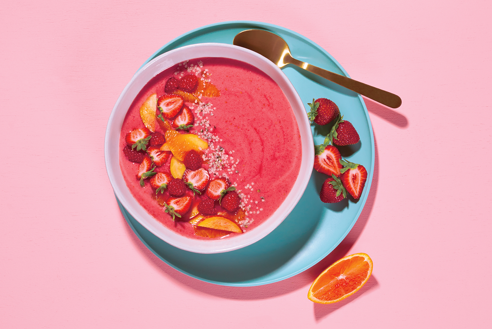 Vibrant pink berry smoothie bowl on a teal plate, garnished with strawberries, raspberries, peach slices, and seeds, accompanied by a gold spoon.