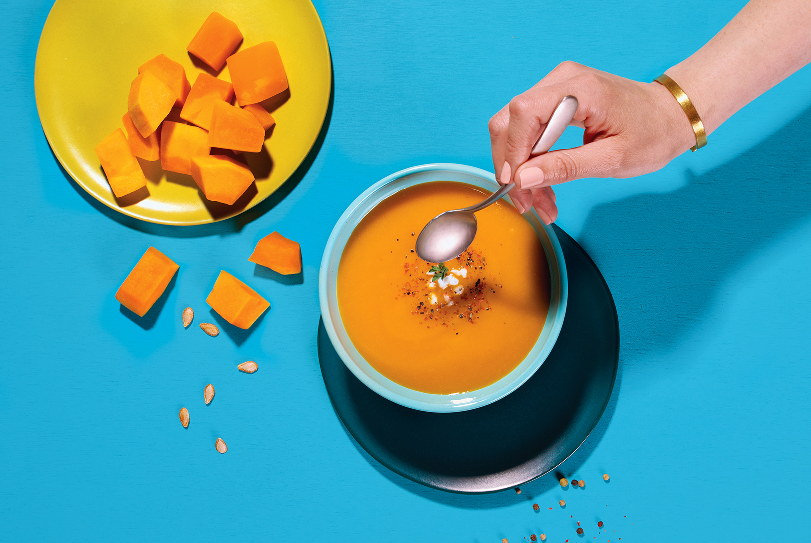 Overhead shot of Butternut Squash and Apple Soup being tasted, alongside diced butternut squash and seeds on a bright blue surface.
