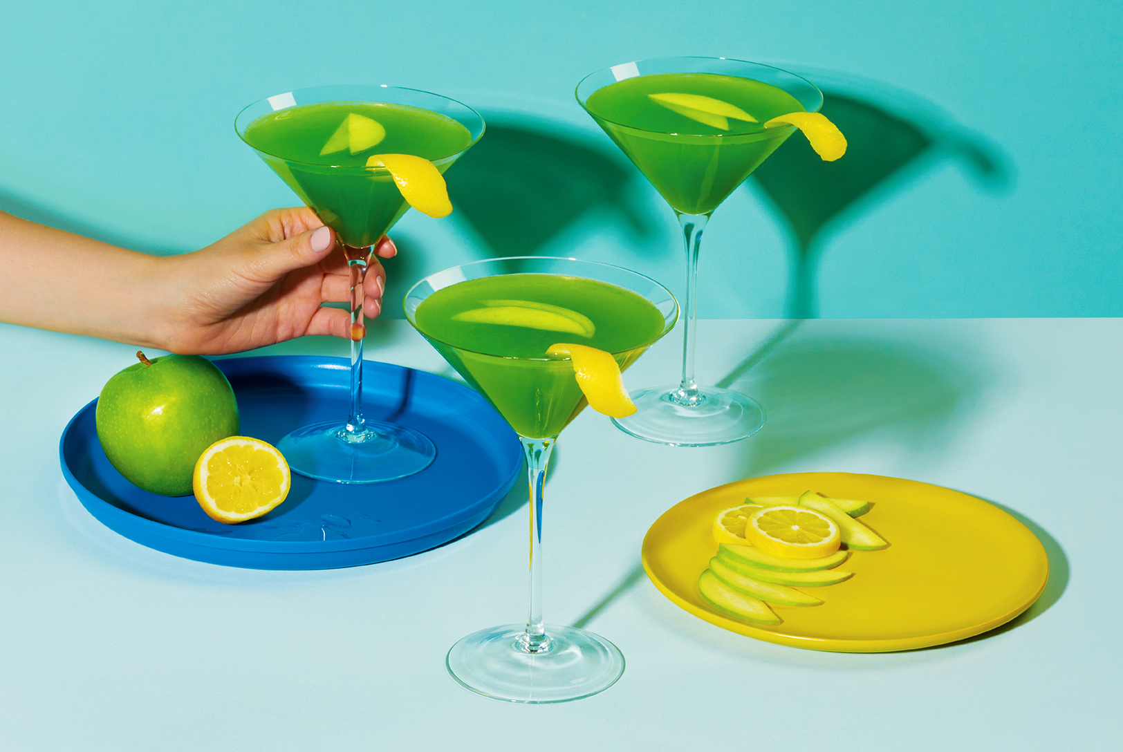 Three vibrant Green Apple Martinis in martini glasses, garnished with lemon twists and apple slices, with fresh fruit nearby.